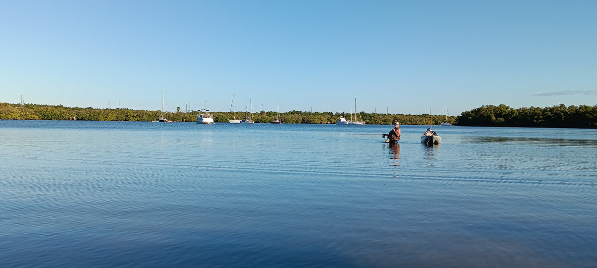 Guide Gary wades waist-deep through calm shallows beside his supply kayak on a guided wade fishing trip in St. Pete, FL, with a mangrove shoreline and anchored sailboats in the background.
