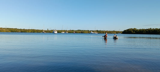 Guide Gary wades waist-deep through calm shallows beside his supply kayak on a guided wade fishing trip in St. Pete, FL, with a mangrove shoreline and anchored sailboats in the background.