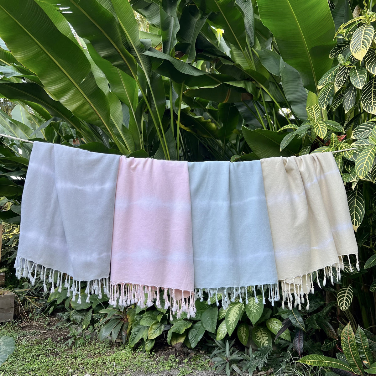 Four pastel tie-dye Turkish towels in gray, pink, blue, and cream hanging on a line against tropical foliage