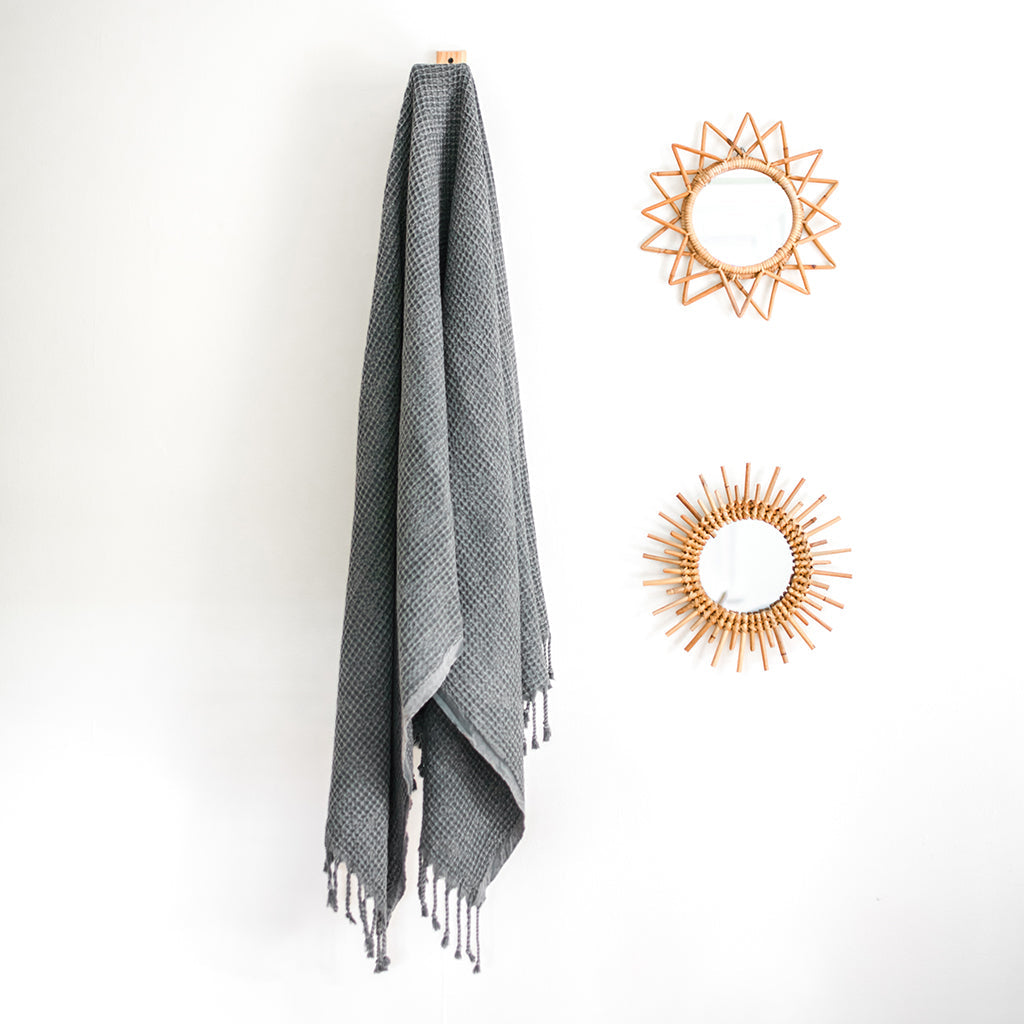 Gray waffle weave Turkish towel hanging on a wall hook beside rattan sunburst mirrors